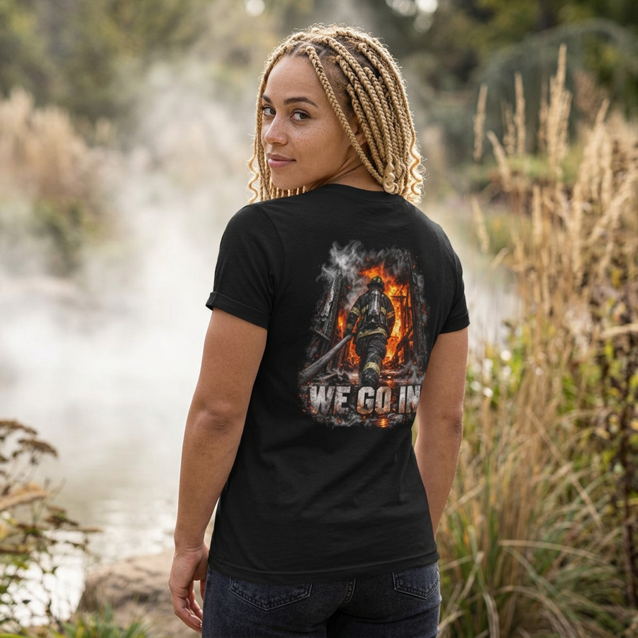 Woman in a black Into the Fire firefighter shirt, back view with a firefighter walking into flames above WE GO IN